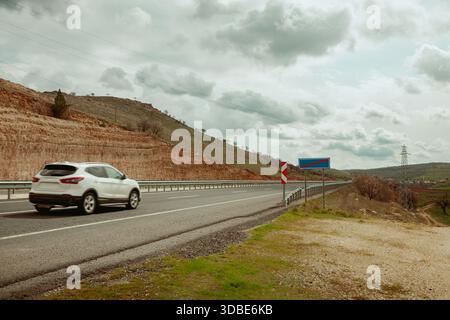 An asphalt road that passes through beautiful lands under a cloudy sky shows the concept of traveling. A white SUV car travels on a countryside road. Stock Photo