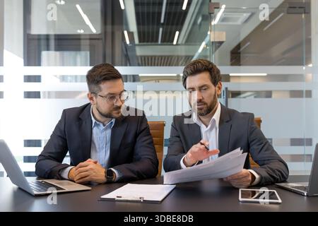Three businessmen in a modern conference room analyze financial reports and strategize together over laptops, tablets and paperwork, collaborating on corporate planning and data review Stock Photo
