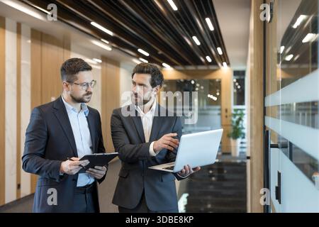 Two professional businessmen collaborating and sharing ideas, one holding a laptop while the other holds a clipboard, discussing important business matters in a modern office hallway Stock Photo