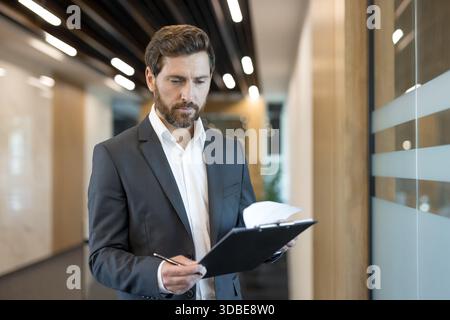 Focused businessman in a suit standing in a modern office corridor, diligently examining documents on his clipboard, reflecting dedication and professional commitment to his work Stock Photo
