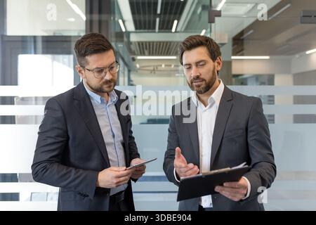 Two professional businessmen collaborating and analyzing documents in a modern office, engaging in a focused discussion about strategic business development and partnership Stock Photo