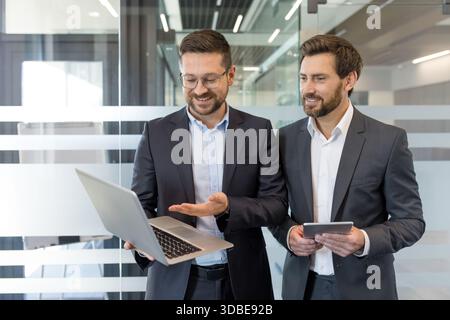 Two smiling businessmen collaborating in a bright office environment, sharing ideas and reviewing information on a laptop and digital tablet, highlighting teamwork and modern communication Stock Photo