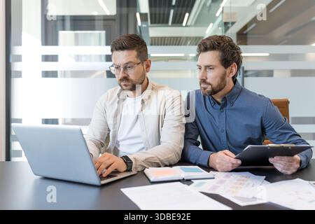 Two business colleagues working together, analyzing data on a laptop and reviewing documents, demonstrating teamwork and professional collaboration in a modern office environment Stock Photo