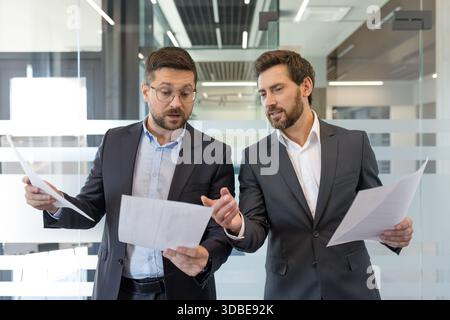 Two business colleagues standing, reviewing and discussing important project documents in a corporate office setting, collaborating on strategy and decision-making Stock Photo