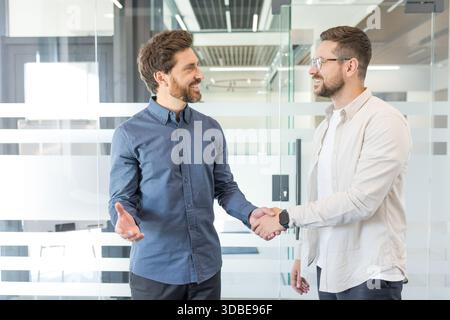 Two smiling adult businessmen standing in a modern office, shaking hands and discussing a successful collaboration and partnership during a professional business meeting Stock Photo