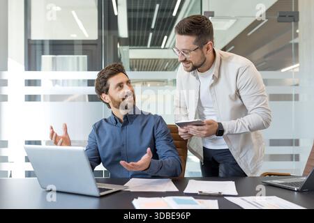 Two business partners are discussing ideas and strategy, smiling and collaborating on a project with a laptop and digital tablet in a modern office environment Stock Photo
