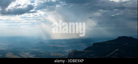 Aerial view of sun rays piercing through dark clouds over the undulating landscape, casting shadows on the hills, Nashik, Maharashtra, India. Stock Photo