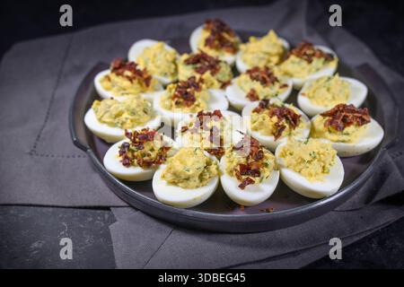 Halved eggs stuffed with a creme from yolk, mustard and herbs and garnished with crispy bacon, homemade party snack on a plate and a dark napkin, copy Stock Photo