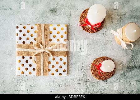 Holiday flat lay with white candles and a rustic gift wrapped in polka dot gold paper and burlap ribbon. Cozy and minimal festive setup Stock Photo