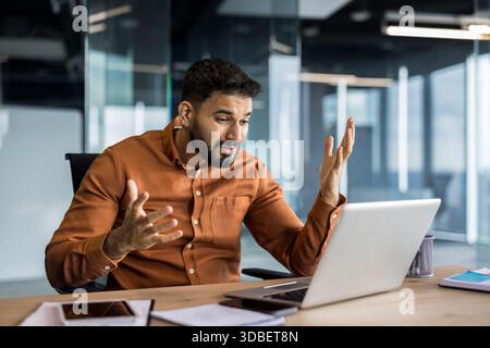 Young businessman actively participating in a video call, talking and gesturing with hands while using a laptop at his office desk during an online meeting or presentation Stock Photo