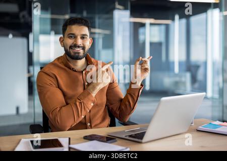 Smiling indian businessman in a brown shirt sits at a desk with laptop, pointing confidently toward copy space in a bright modern office, suggesting guidance and recommendation Stock Photo