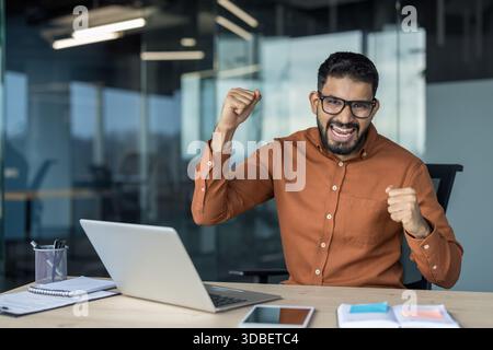 Excited young indian man celebrating good news or achievement while sitting at his desk, working on a laptop in a contemporary office setting, showing joy and victory Stock Photo