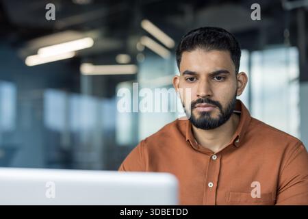 Indian businessman with a beard, serious and focused at his laptop in a modern office, concentrating on tasks and managing business work in a professional startup environment Stock Photo