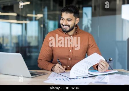 Smiling indian businessman working at his desk, reviewing paperwork and using a laptop in a modern office, representing professional success, administration, and efficient financial management Stock Photo