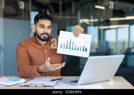 Man holding and presenting a bar chart to colleagues during a remote video conference, discussing business data and financial statistics in a modern office setup Stock Photo
