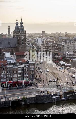 View of historic buildings with red awnings and a bustling street with cars and trams under a soft sky, Amsterdam, North Holland, Netherlands. Stock Photo