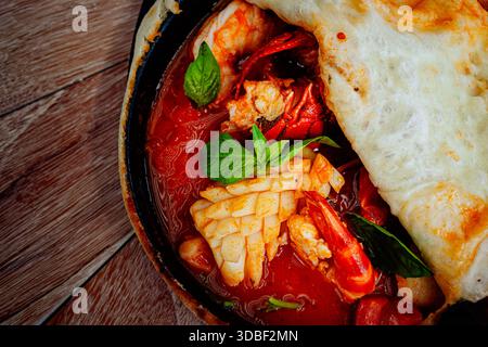 Seafood stew en croute simmers in a cast iron pan on a weathered table, garnished with basil leaves and crusty bread. The rich stew features succulent Stock Photo