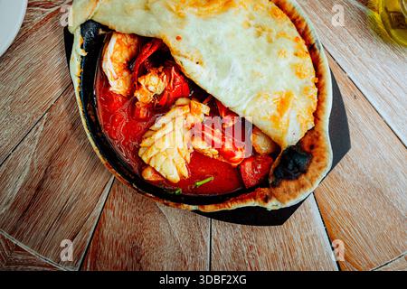 Seafood stew en croute features shrimp, squid, and crab in a cast iron skillet covered with bread on a wooden table beside oil. Vibrant red peppers ad Stock Photo