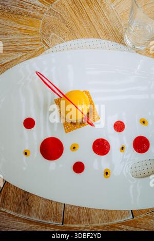 Mango sorbet dessert with red fruit coulis dots layered on crisp pastry sits on a white ceramic plate, accompanied by water and a red plastic fork ato Stock Photo
