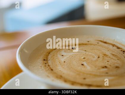 A white ceramic cup filled with dark espresso and textured foam sits on a white saucer atop a grainy wooden tabletop near a blue-painted window. Stock Photo
