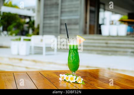 A vibrant tropical drink sits on a glossy wooden table adorned with yellow and white flowers. A pink fruit slice and ice cubes complement the green li Stock Photo