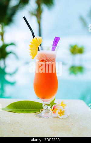 A vibrant tropical cocktail in curved glass, garnished with pineapple wedge, straw, and umbrella sits against a backdrop of glistening pool water, san Stock Photo