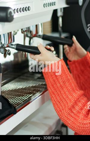 Female hands in vibrant knit sweater pulling levers on professional coffee machine in cafe setting Stock Photo