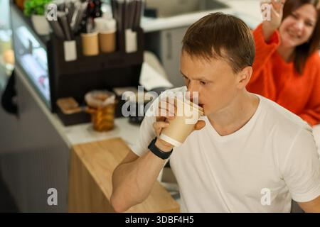 Coffee shop moment: man in white shirt drinks from takeaway cup, woman in orange sweater waves enthusiastically in background. Stock Photo