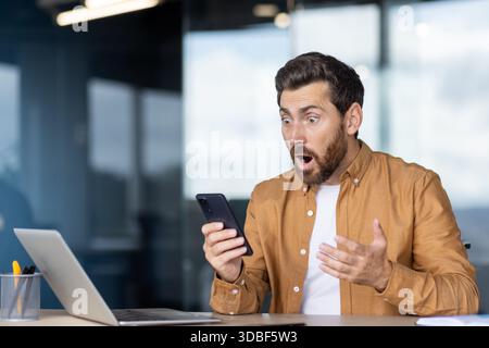 Surprised businessman reading unexpected news on smartphone at office desk, wide eyed and mouth agape while checking laptop, reacting to shocking online message or alert Stock Photo