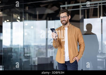 Man smiling while interacting with a smartphone, engaging in digital communication or mobile business activities within a clear glass office environment Stock Photo