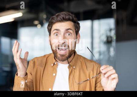 Man with beard and brown hair showing a wide-eyed expression of surprise and excitement, holding eyeglasses in hand, reacting to unexpected news or discovery in a modern office setting Stock Photo