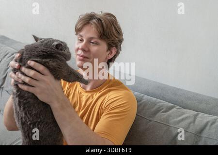 Holding cat and looking at it with care, young man spending quiet time with his pet at home on sofa. Man Spending Time with His Cat at Home Stock Photo