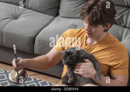 Holding mirror while sitting on floor, young caucasian man spends time with his pet cat in calm home environment. Stock Photo