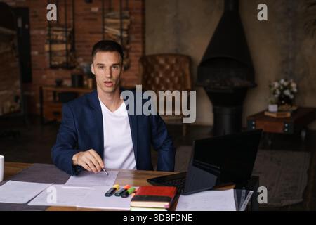 Young man in jacket working from home doing paperwork using laptop and holding a pen in his hand while sitting at his desk. Performs work remotely || Model released Stock Photo
