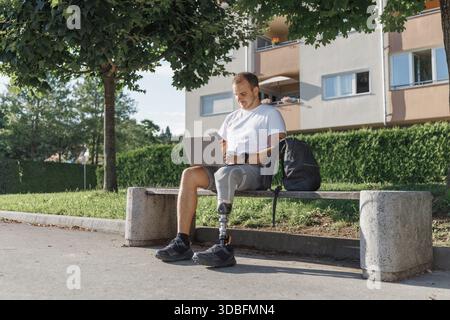 Man with a prosthetic leg using wireless technology devices while sitting outdoors on a bench, working on a laptop and talking on a smartphone. Stock Photo