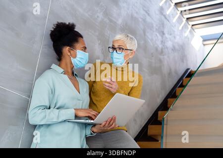 Female professionals in masks using laptop for technology, AI, digital innovation and safe workplace Stock Photo