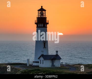The solitary Heceta Head Lighthouse stands on a grassy cliff overlooking the ocean. The sun sets in the background, casting an orange glow across the Stock Photo