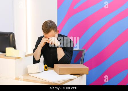 Man in black shirt sits rubbing face at desk with packages, tape, papers, colorful background. Stock Photo