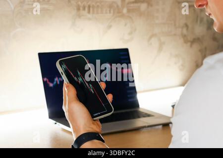 Profile of person examining downward stock trend on smartphone with open computer showing similar data Stock Photo