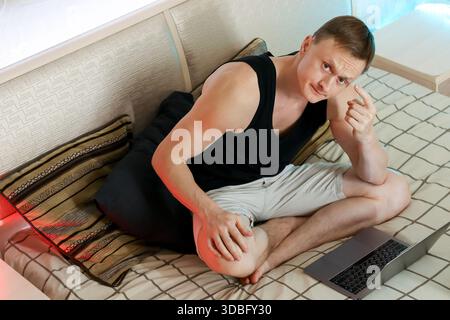 Young male in casual attire makes hand gesture while sitting beside open laptop on bed in home. Stock Photo