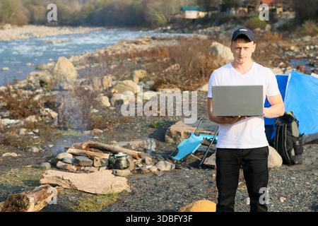 Profile of a man in cap and white v-neck shirt gripping a laptop, with blue tent, backpack, and smoky fire in the background on autumn riverside. Stock Photo