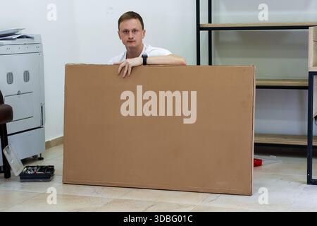 Man standing behind large cardboard sheet looking over. Man in casual wear standing and resting hand on top of upright large cardboard panel in room w Stock Photo