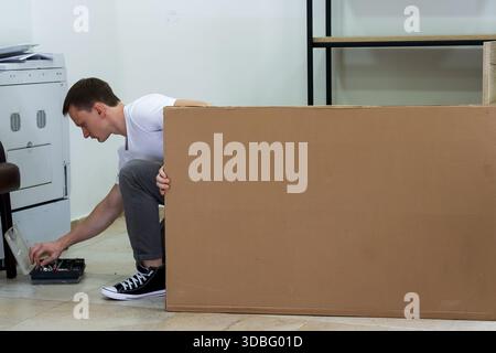 Man crouching and reaching for tools while hiding behind cardboard. Young man in casual clothes crouching behind large cardboard sheet reaching for sc Stock Photo