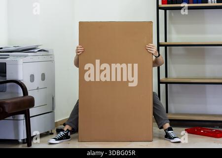 Person sitting holding bottom of large furniture cardboard box. Seated figure grasping lower edges of tall flat-pack furniture box concealing body in Stock Photo