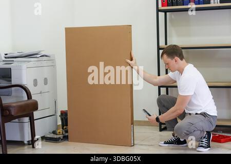 Man crouching next to large cardboard package holding smartphone. Adult male in white t-shirt crouching and touching big cardboard box while looking a Stock Photo