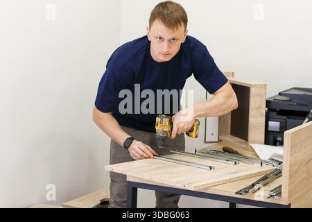 Carpenter drilling holes for drawer slides in furniture. Man precisely drilling into wood panel to mount metal drawer guides during furniture build Stock Photo