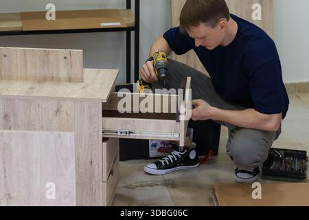 Man using cordless drill to screw drawer slide in cabinet. Man crouching and using cordless drill to fasten metal drawer slide inside light wood cabin Stock Photo