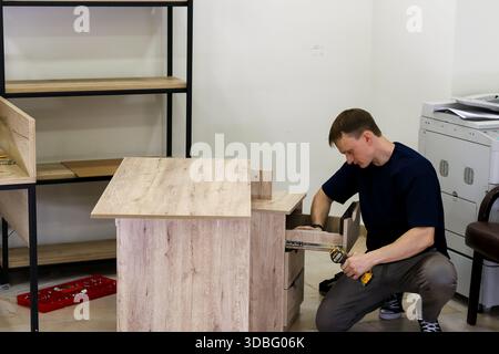 Worker assembling modern wooden cabinet with tools. Man focused on attaching drawer slides to light oak cabinet using cordless screwdriver in workshop Stock Photo