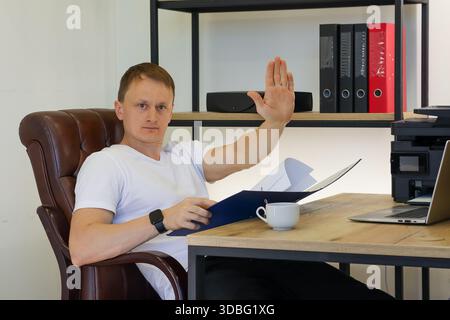 Man in office waves hand upwards in greeting or signal while reviewing open blue folder on wooden desk with tech. Stock Photo