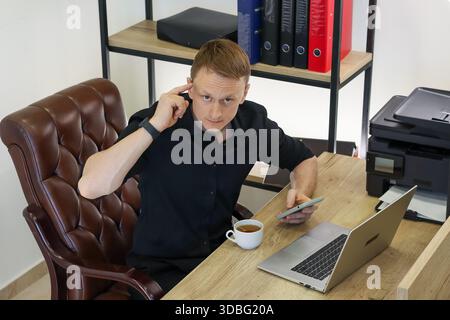 Professional seated at desk holds mobile, reaches to ear with finger, with computer and hot beverage in view. Stock Photo
